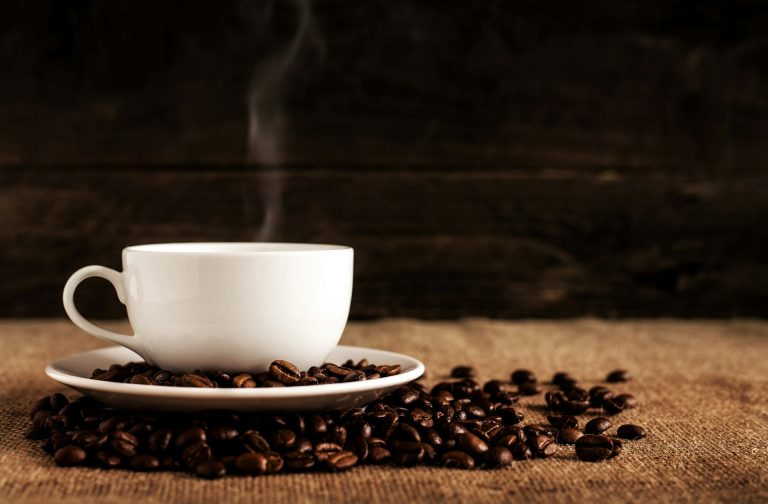 cangkir keramik putih dengan biji kopi di atas kain coklat bertekstur